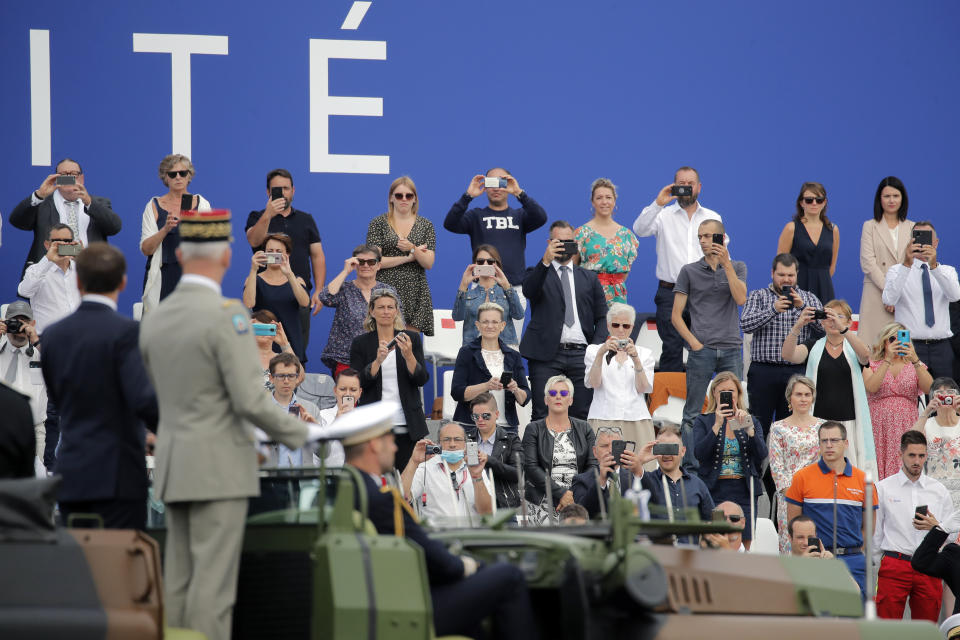 People take snapshots of France's President Emmanuel Macron, foreground left, and French Armies Chief Staff General Francois Lecointre, foregound second from left, as they stand in the command car to review troops before the start of the Bastille Day military parade, Tuesday, July 14, 2020 in Paris. France are honoring nurses, ambulance drivers, supermarket cashiers and others on its biggest national holiday Tuesday. Bastille Day's usual grandiose military parade in Paris is being redesigned this year to celebrate heroes of the coronavirus pandemic. (AP Photo/Christophe Ena, Pool)