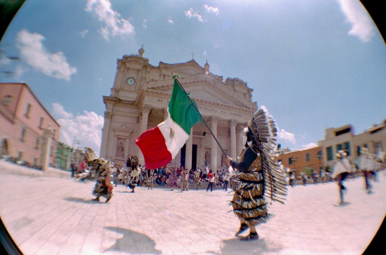 Learn more about Mexican culture and traditions with these 10 fun facts about Mexico: Pictured: a distorted image of person in traditionally clothing holding a Mexican flag in front of a building.