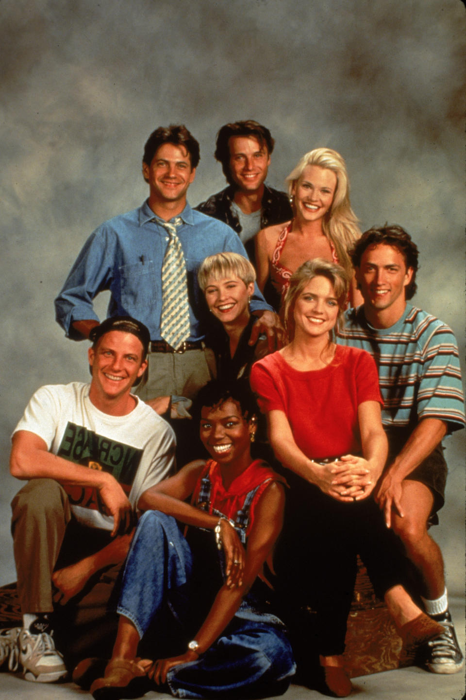 The main cast of Melrose Place stand and sit together smiling in a promotional shot from the early 90s