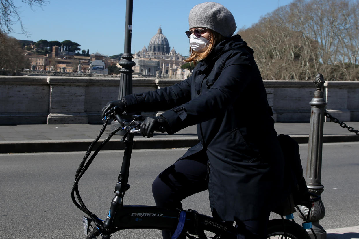 ROME, ITALY - MARCH 11: A girl wearing a protective mask crosses a bridge by bicycle in front St. Peter's Basilica on March 11, 2020 in Rome, Italy. The Italian Government has taken the unprecedented measure of a nationwide lockdown in an effort to fight the world's second-most deadly coronavirus outbreak outside of China. The movements in and out are allowed only for work and health reasons proven by a medical certificate. The justifications for the movements needs to be certified with a self-declaration by filling in forms provided by the police forces in charge of the checks. (Photo by Franco Origlia/Getty Images)