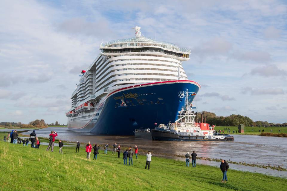 The Carnival Jubilee is transported by tugboat while a crowd watches from the grassy shore.