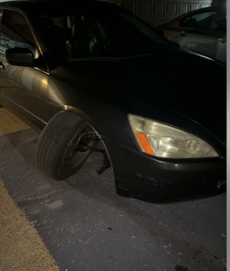 A black car with a severely damaged front left wheel is parked on a dark surface. The wheel is bent inward, rendering the car unable to drive