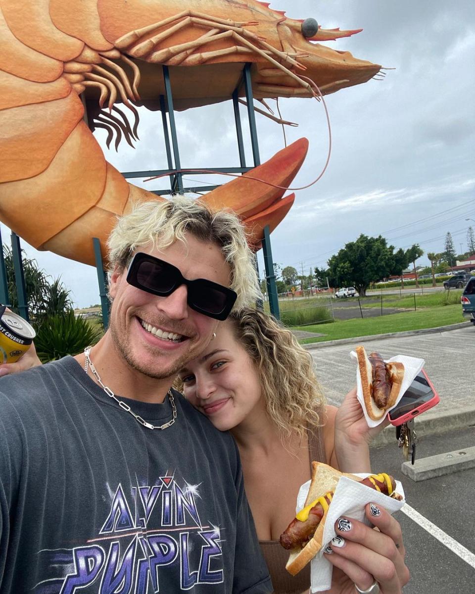 The Bachelor's Abbie Chatfield and boyfriend Konrad Bien Stephens pose for a photo in front of a giant prawn statue while holding sausages in bread. Photo: Instagram/abbiechatfield.