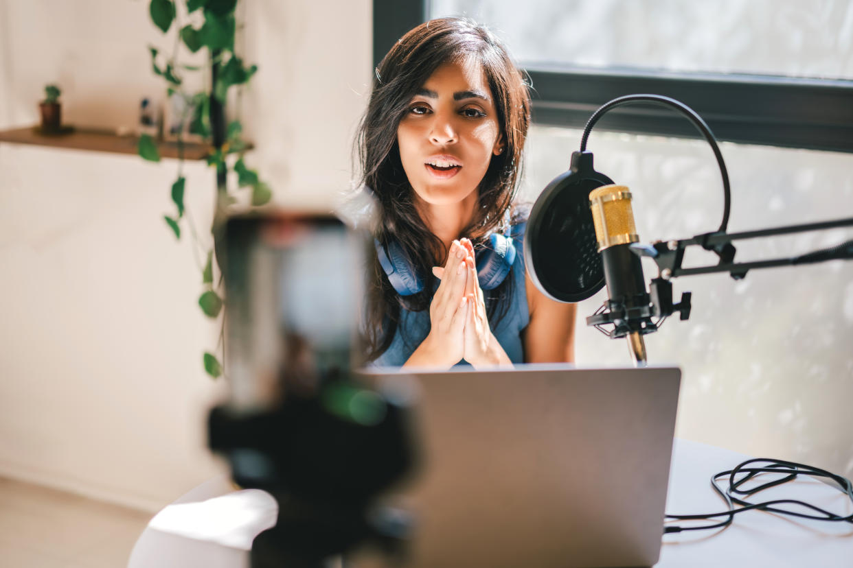 A beautiful Asian businesswoman is recording a vertical video for her social media online course, talking about her expertise and life story, and she is using a laptop, studio microphone, and a smart phone on a tripod. Clean and minimalistic domestic set up interior with plants and natural lighting. Professional creativity and authenticity