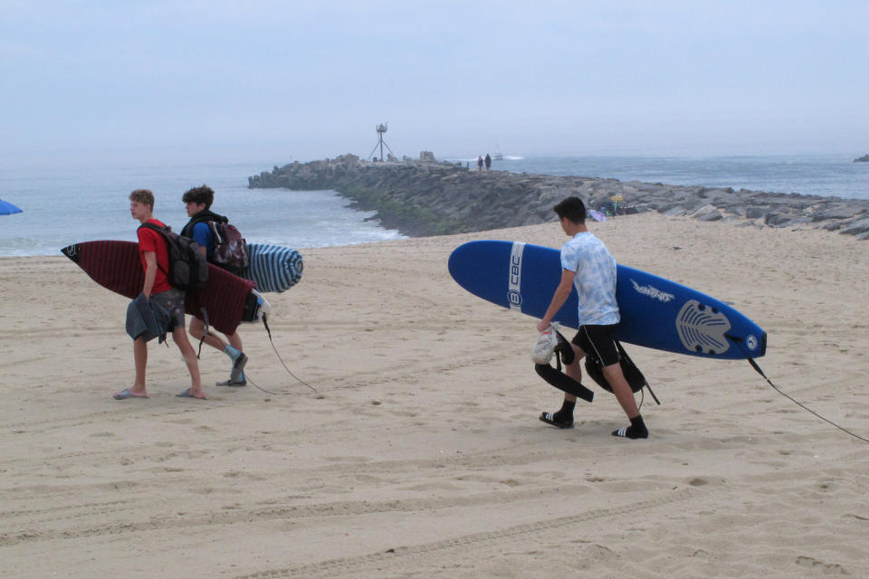 In this June 17, 2019 photo, surfers carry their boards across the beach in Manasquan N.J. where there used to be sand dunes. The town decided not to rebuild its dunes after Superstorm Sandy in 2012, but the U.S. Army Corps of Engineers will review whether the handful of places along the New Jersey shore that don't have dunes now should build them. (AP Photo/Wayne Parry)