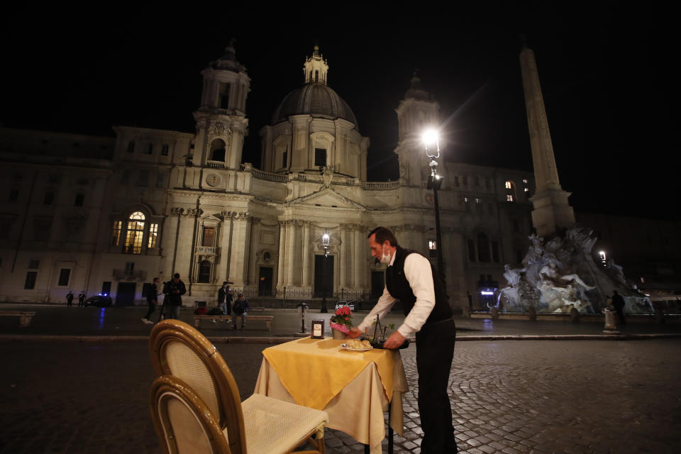 A waiter clears a table at a cafe restaurant in Piazza Navona Square before the start of a curfew, Friday, Oct. 23, 2020. In much of Europe, city squares and streets, be they wide, elegant boulevards like in Paris or cobblestoned alleys in Rome, serve as animated evening extensions of drawing rooms and living rooms. As Coronavirus restrictions once again put limitations on how we live and socialize, AP photographers across Europe delivered a snapshot of how Friday evening, the gateway to the weekend, looks and feels. (AP Photo/Alessandra Tarantino)