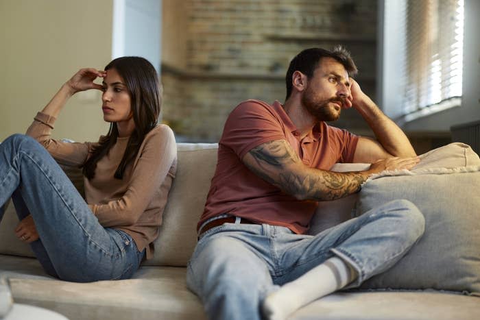 Two people sit on a couch, turned away from each other, appearing upset. Their body language suggests they might be in disagreement or deep thought