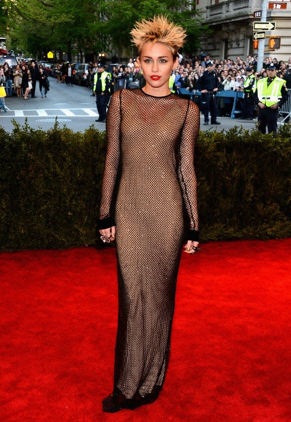 Miley Cyrus poses on the red carpet wearing a floor-length fishnet dress, and her short hair styled in spikes.