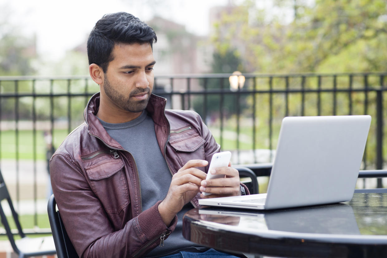 Man in burgundy leather jacket sitting outside on his laptop using his phone.