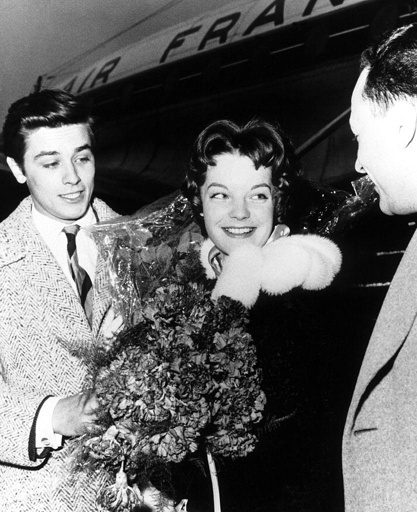 C’est donc de bonne grâce que le divin Alain, alors au faîte de sa beauté du haut de ses 23 printemps, a accepté d’aller chercher Romy Schneider à l’aéroport, un bouquet de roses à la main - une idée de la production du film, désireuse de faire de belles images de promotion. En le voyant sur le tarmac, la jeune actrice n’est pourtant pas plus émue que cela par l’intensité ombrageuse du regard du jeune premier. Bien au contraire : "<em>Au pied de l’escalier roulant, je découvris un jeune type beaucoup trop beau, beaucoup trop bien coiffé, en cravate, dans un costume trop à la mode…</em>", dira-t-elle plus tard, amusée. (<em>Keystone-France/Gamma-Keystone via Getty Images</em>)