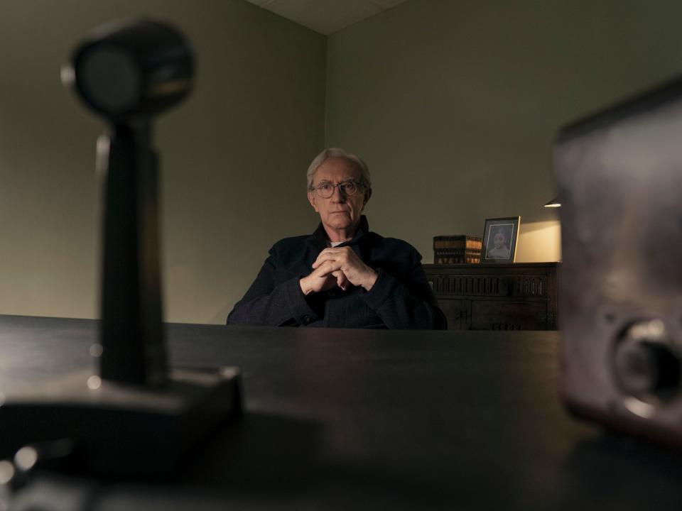 mike evans in 3 body problem, sitting at a desk with an old school microphone and transmitter box on it. there's a shelf behind him with a photo of someone, and a small box