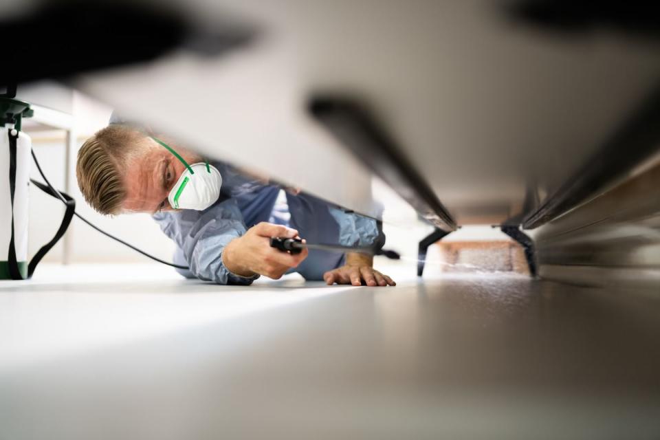 An exterminator wearing a mask applies a product under a piece of furniture. 