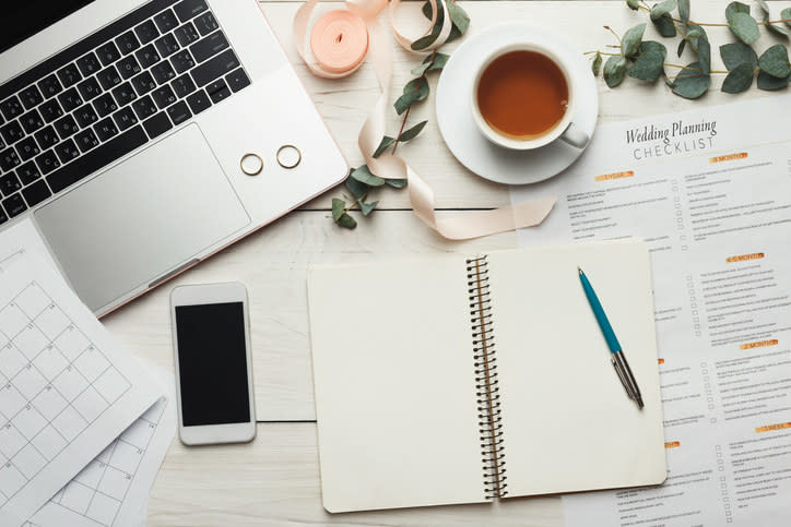 A notebook opened next to a cellphone, computer, cup of tea, and wedding planning checklist.