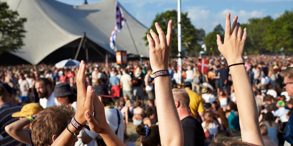 Stock image of two friends raising their hands, wearing festival bracelets, at an outdoor festival, crowds are sitting down blurred in the background, a tent is in the far background.