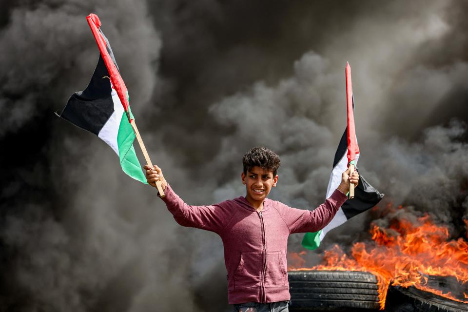 A youth holds Palestinian flags as he stands by the fumes from flaming tires during a demonstration along the border with Israel east of Gaza City on April 5, 2023. - Israeli police said they had entered to dislodge "agitators" from Jerusalem's al-Aqsa Mosque, a move denounced as an "unprecedented crime" by the Palestinian Islamist movement Hamas. The holy Muslim site is built on top of what Jews call the Temple Mount, Judaism's holiest site. (Photo by MAHMUD HAMS / AFP) (Photo by MAHMUD HAMS/AFP via Getty Images) ORG XMIT: 4978 ORIG FILE ID: AFP_33CP432.jpg