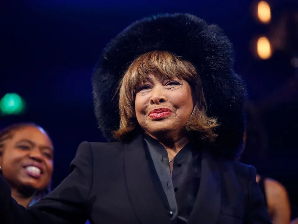 Tina Turner standing onstage with the cast of &quot;Tina: The Tina Turner Musical&quot; while wearing a black blazer, black shirt, and black hat.