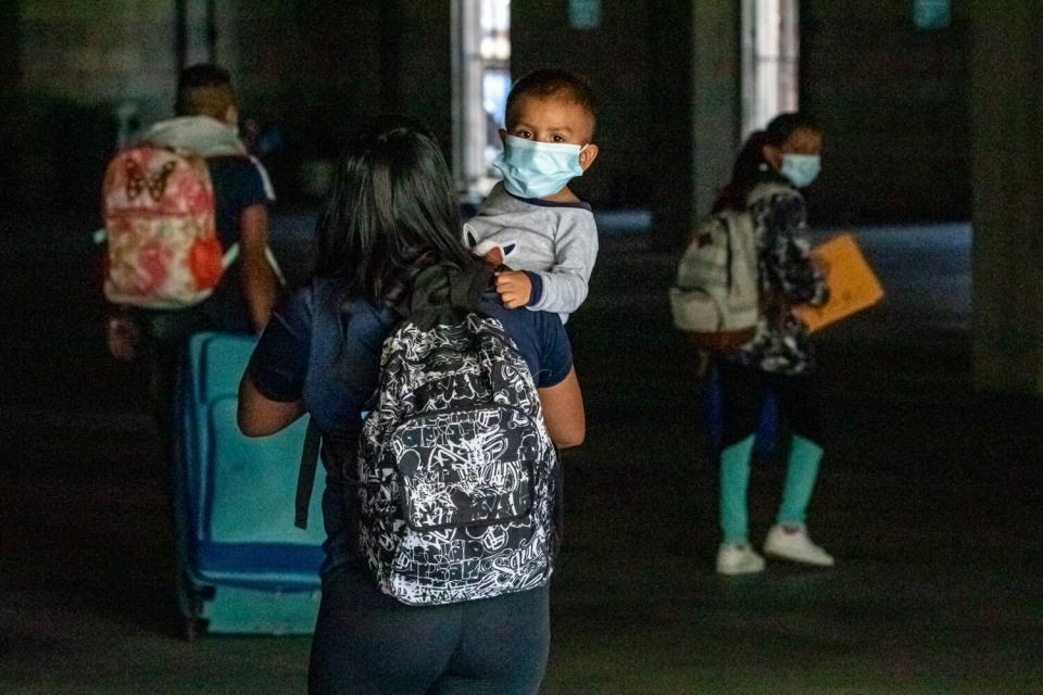 A woman with a backpack carries a small child as other people with bags walk nearby