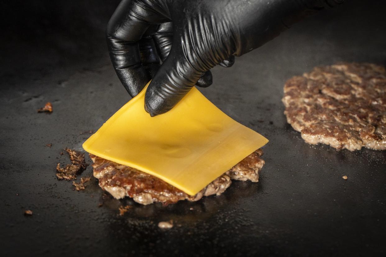 burger patty on griddle, gloved hand placing slice of cheese on top