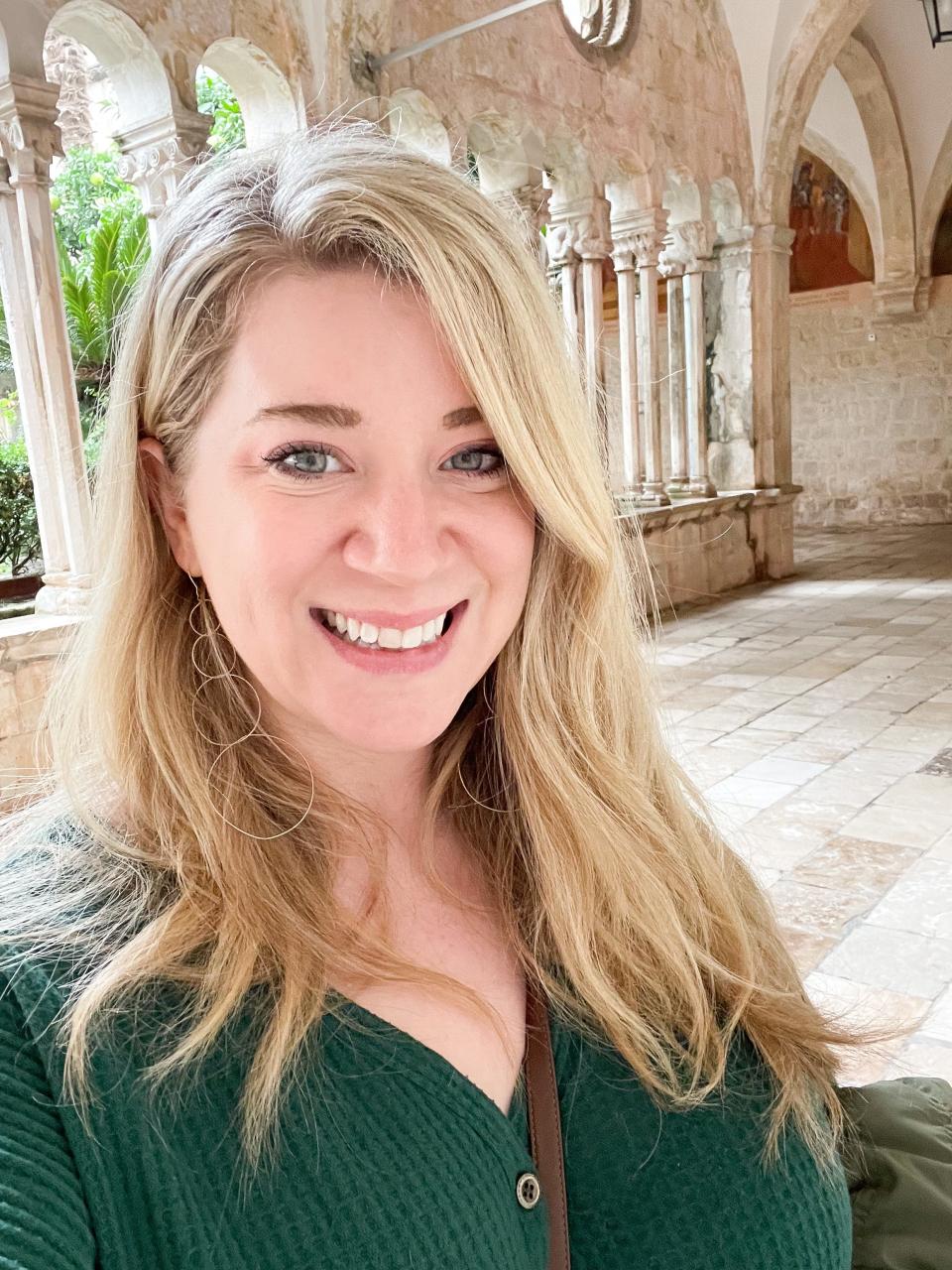 A blonde woman smiling at the camera in front of arched stone walkway.