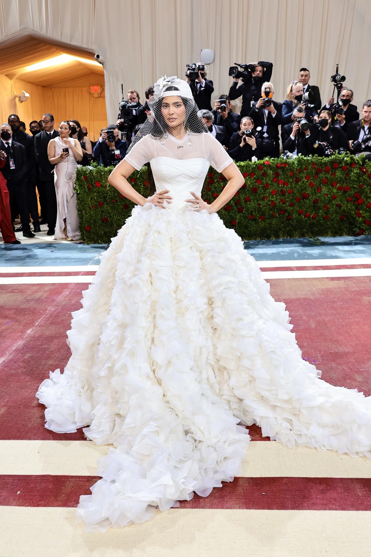 Kylie Jenner attends the 2022 Met Gala in a bridal-inspired look: A dress with a large white skirt, a sheer t-shirt style top, and a white baseball cap with a white birdcage veil.