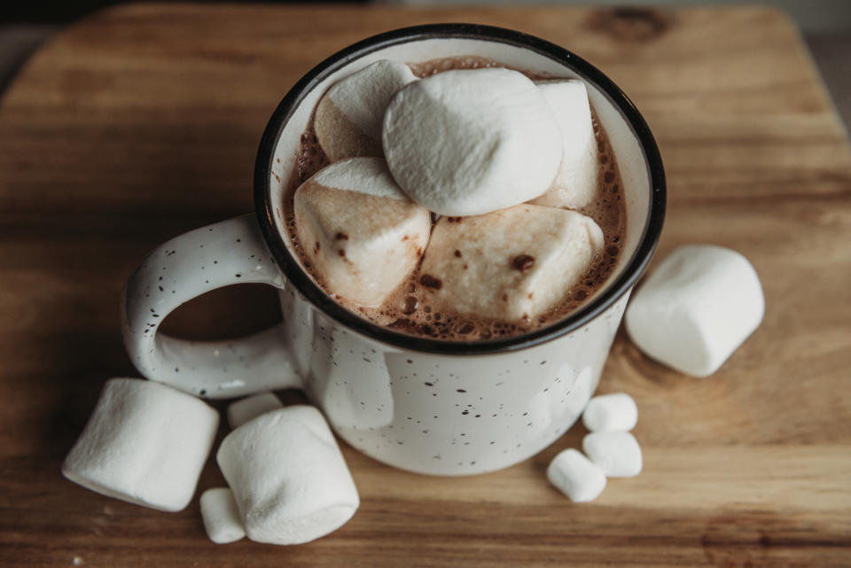 A ceramic mug filled with hot chocolate topped with marshmallows sits on a wooden surface, with additional marshmallows scattered around it