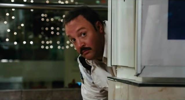 Paul Blart is looking out from the side of the wall to see what is happening in front of him