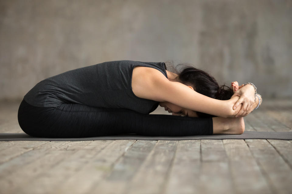 Young yogi woman practicing yoga, doing paschimottanasana exercise, Seated forward bend pose, working out, wearing sportswear, black pants and top, indoor full length, gray wall in yoga studio