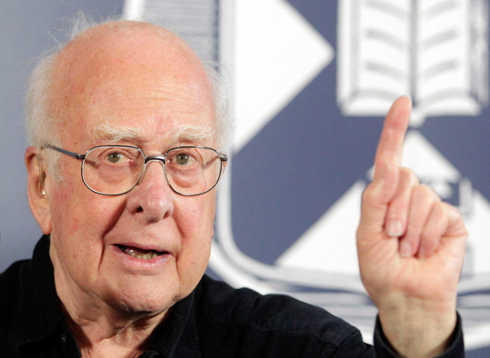 A close up of a man with white hair and glasses. He's raising his left hand and has one finger pointing up.