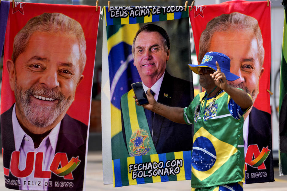 ARCHIVO - Un manifestante vestido con los colores de la bandera brasileña frente a un puesto de toallas que muestran a los candidatos presidenciales, el actual mandatario Jair Bolsonaro (centro) y al expresidente Luiz Inácio Lula da Silva, en Brasilia, Brasil, el 27 de septiembre de 2022. (AP Foto/Eraldo Peres, Archivo)