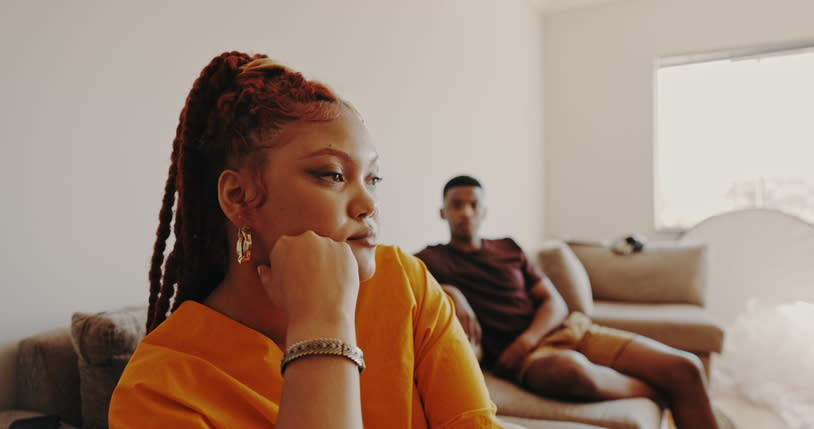 I don't know who the individuals in the image are. The image shows a woman in a pensive pose with a man sitting on a couch in a room, suggesting a relationship issue