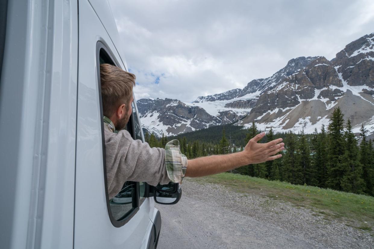 Cheerful young man outstretches arms out of the window of a motor home. Mountain and lake landscape.