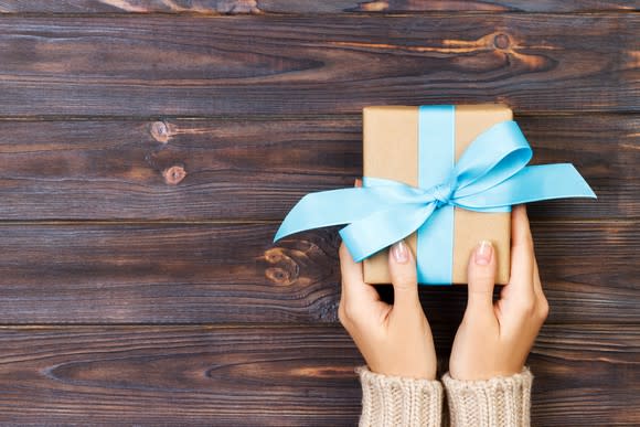 Two hands holding a small wrapped present with blue ribbon on a wood table.