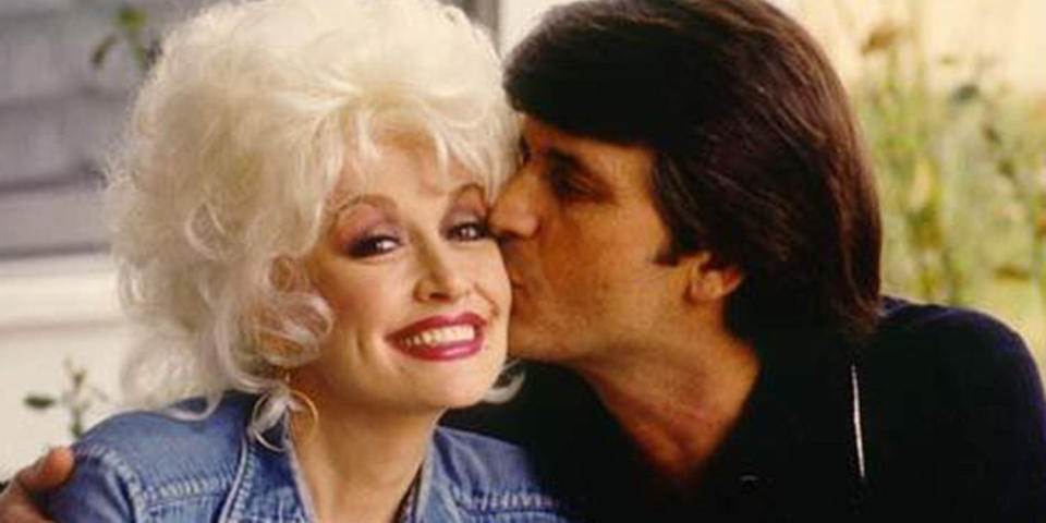 Dolly parton smiles as a dark haired main kisses her cheek. (dollyparton.com)