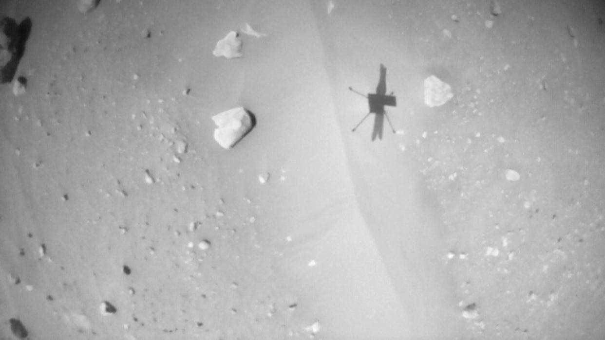  a black-and-white image showing a small helicopter's shadow on sandy, boulder-studded ground. 