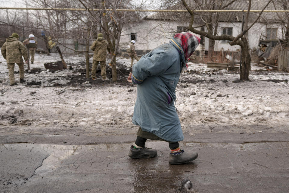 An elderly lady walks by as members of the Joint Centre for Control and Coordination on ceasefire of the demarcation line, or JCCC, survey a crater and damage to a house from artillery shell that landed in Vrubivka, one of the at least eight that hit the village today, according to local officials, in the Luhansk region, eastern Ukraine, Thursday, Feb. 17, 2022. U.S. President Joe Biden warned that Russia could still invade Ukraine within days and Russia expelled the No. 2 diplomat at the U.S. Embassy in Moscow, as tensions flared anew in the worst East-West standoff in decades. (AP Photo/Vadim Ghirda)