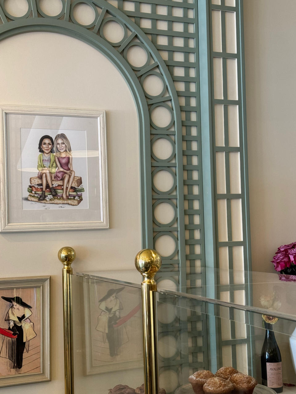 Framed drawing of two children sitting is displayed on a wall in a bakery, with pastries and a bottle of wine on a counter below