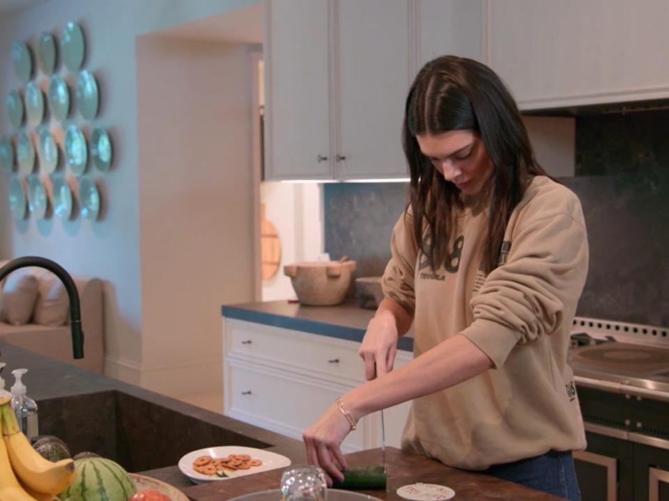 kendall jenner standing in a kitchen trying to cut a cucumber: she's coming at it with a knife in her right hand, but reaching over her body with her left hand to stabilize it