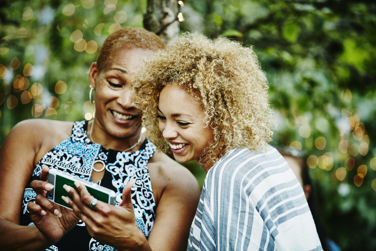 Smiling mature aunt showing niece photos on smartphone during party in backyard of home