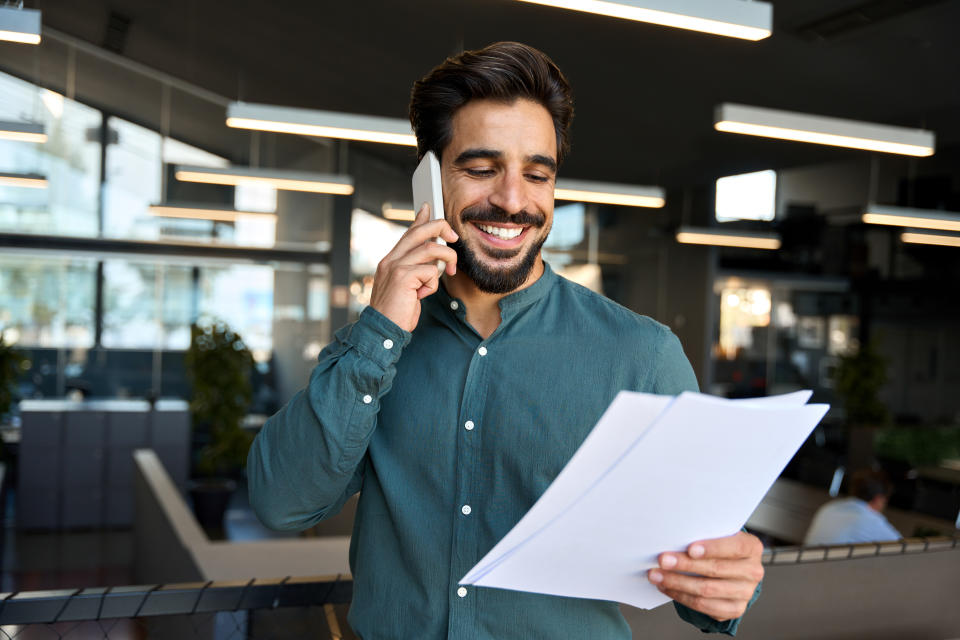 Smiling happy young bearded Latin professional business man executive holding documents and cell phone making mobile call at work on cellphone consulting client standing in modern office.