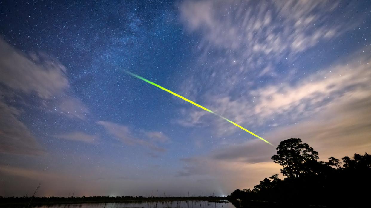  A streak of green light zooms across the night sky above a lake. 
