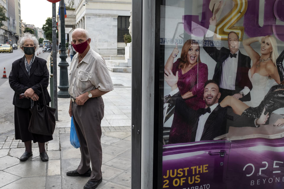 Two people wearing face masks to prevent the spread of COVID-19, wait at a bus station where an advertisement for a TV show is displayed, in Athens, Monday, Nov. 2, 2020. The government announced new lockdown measures Saturday to stem the rapid rise in new cases, among which are the closure of bars, cafes, restaurants and gyms in large swaths of the country, will take effect Tuesday through at least the end of November. (AP Photo/Yorgos Karahalis)