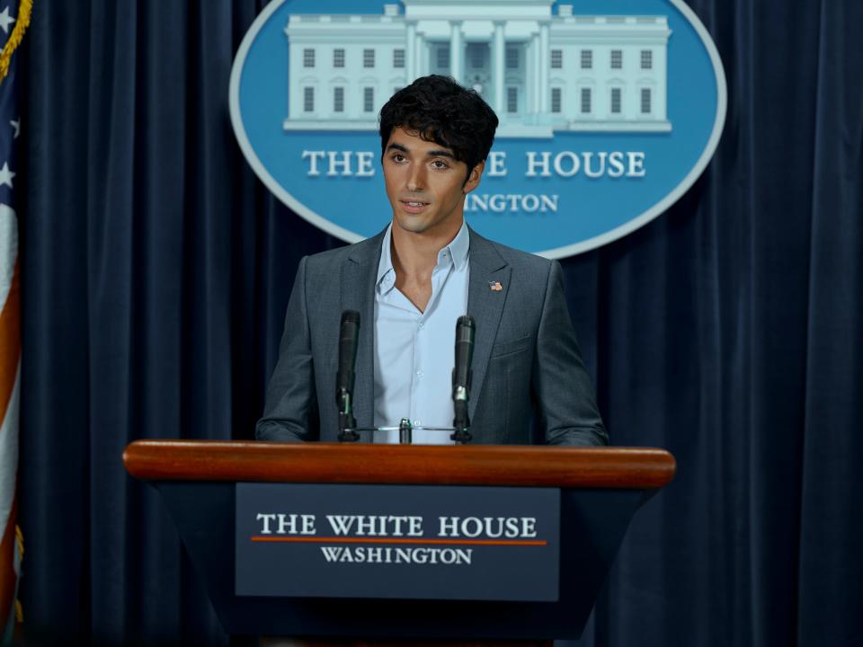 alex in red white and royal blue, dressed in a grey suit and shirt with the top button undone, speaking at a white house podium in the press room