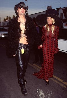 Pamela Anderson wears a long red dress and large black hat, holding hands with ex husband Tommy Lee who is shirtless beneath a feather black jacket and wearing leather trousers.