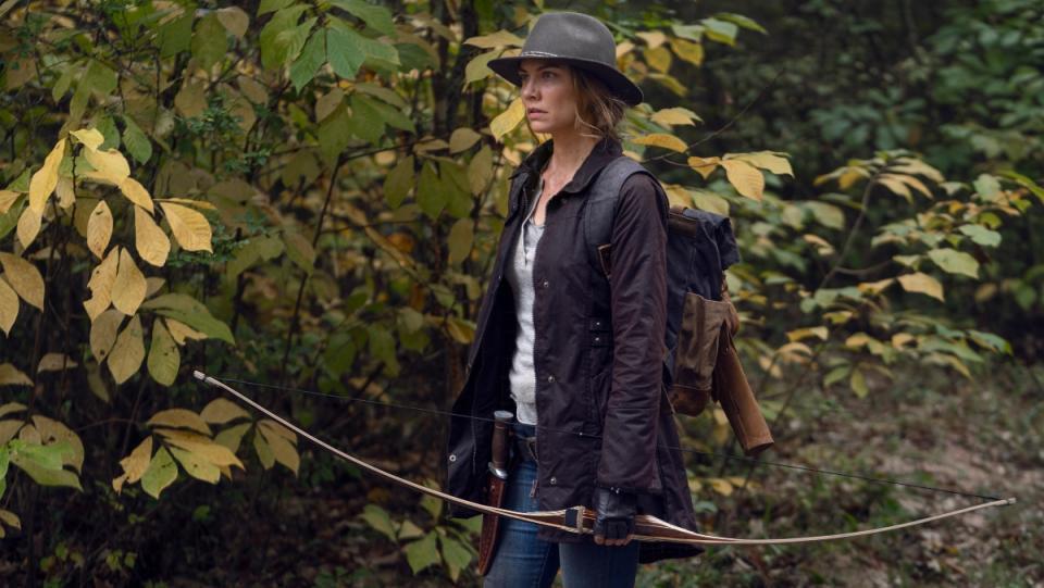 A photo of a woman wearing a brown hat and standing near trees while holding a bow and arrow on The Walking Dead