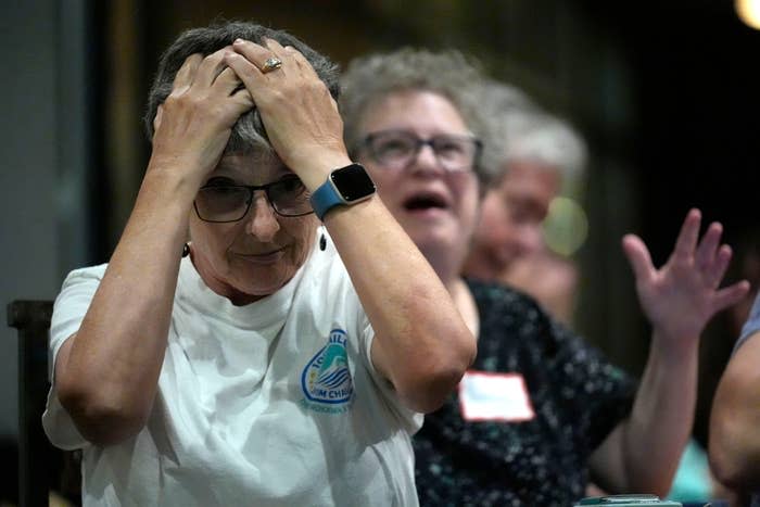 A group of people reacting with surprise or shock. An older woman in glasses and a white shirt holds her head with her hands. Other people appear behind her