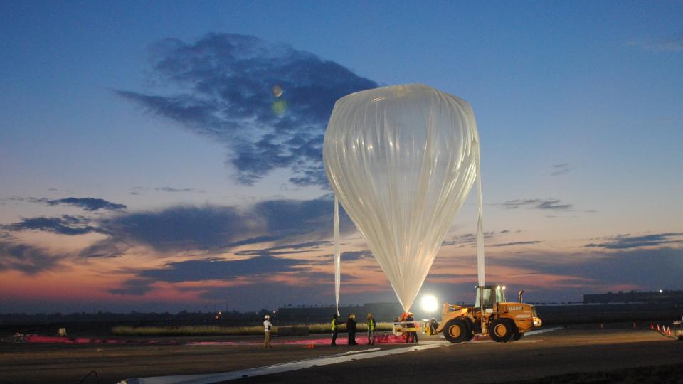 a stratospheric balloon inflating while tied to the ground. a small support vehicle stands at right oft the balloon. in back is a sky at dawn, with orange and blue skies and a few clouds