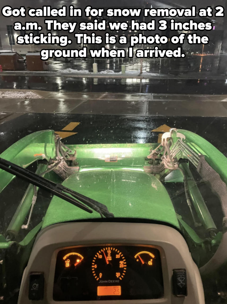 View from inside a John Deere tractor at night, dashboard illuminated, reflecting a lit urban parking lot with wet pavement and surrounding buildings