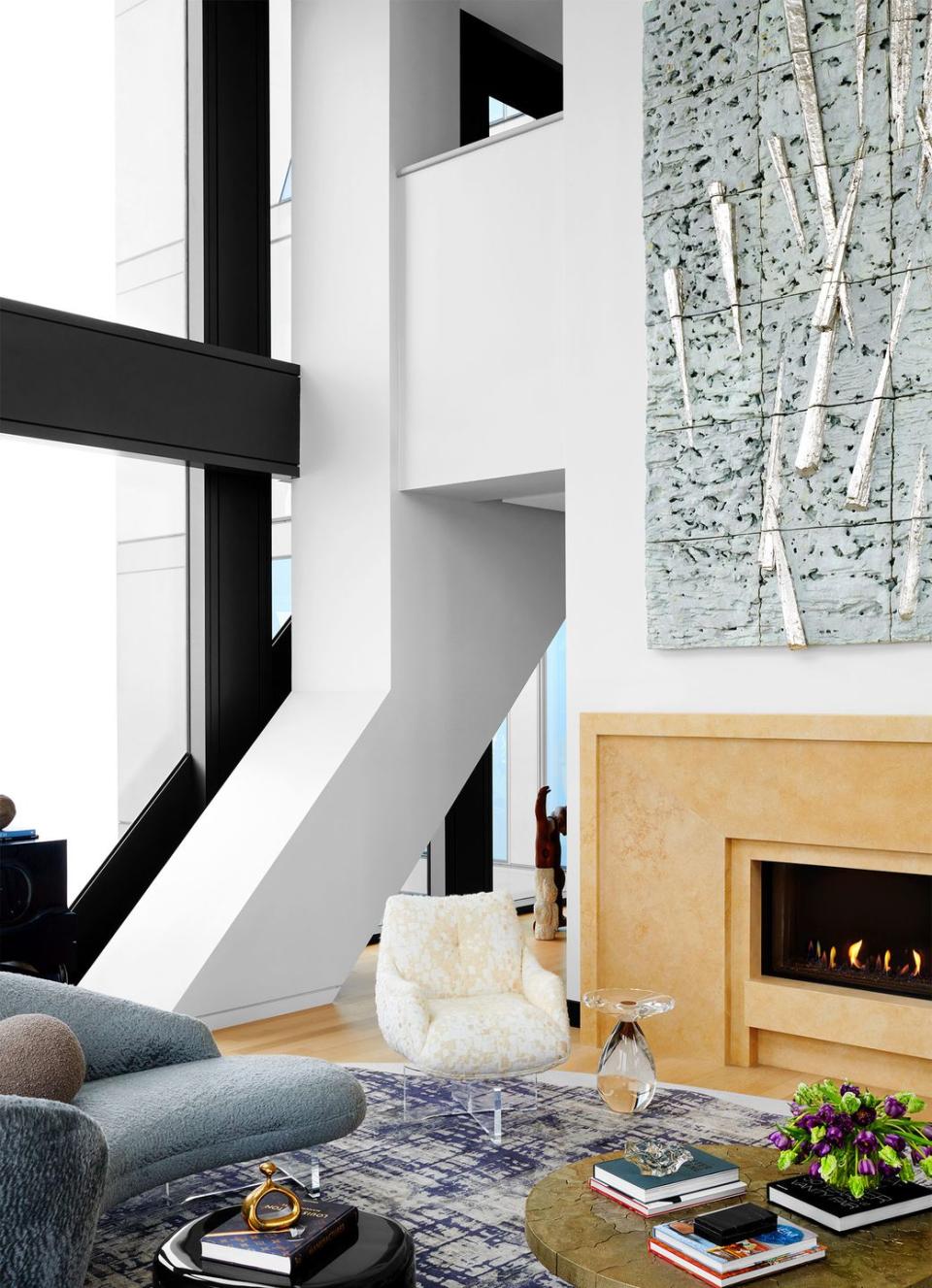 in a living room is a curved sofa in textured blue fabric, accent chair in a light mottled fabric, fireplace, round cocktail table, with books, accent tables, blue and white rug, 3d clay artwork above fireplace