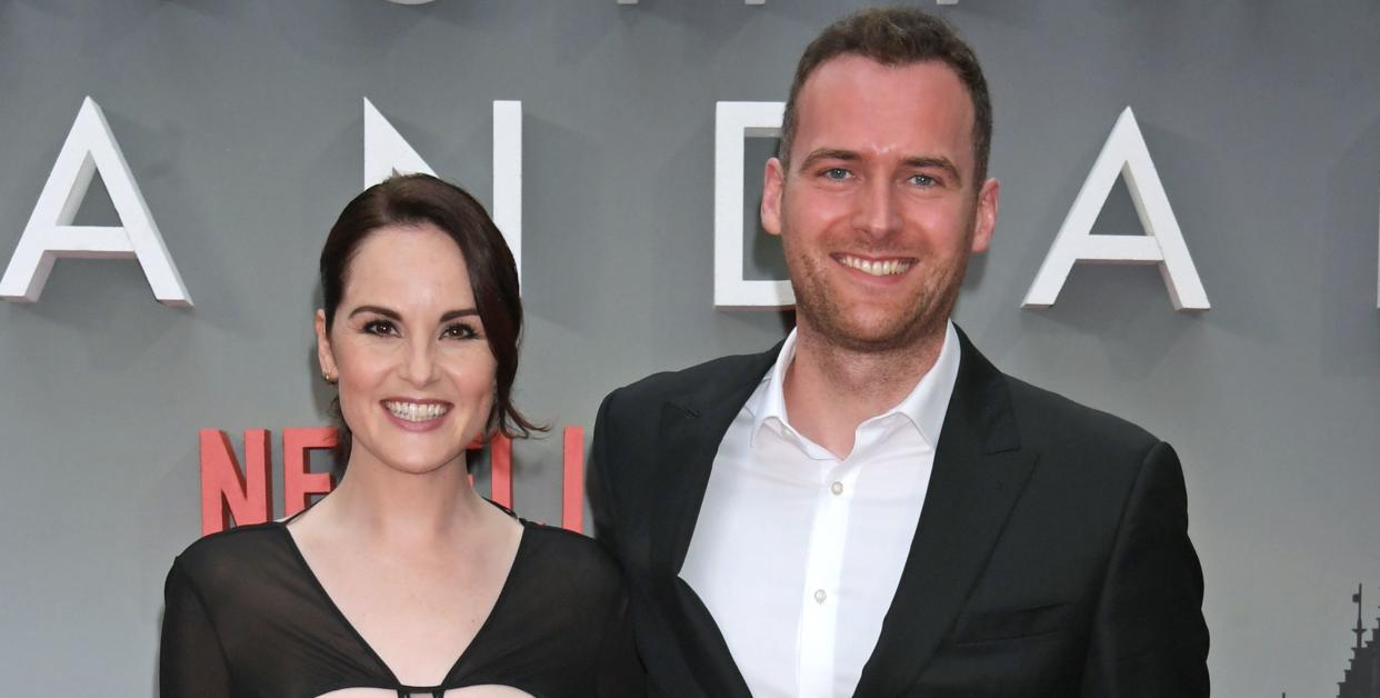 michelle dockery and jasper waller bridge stand with arms around each other and smiling at the camera, she wears a long back dress and he a black suit