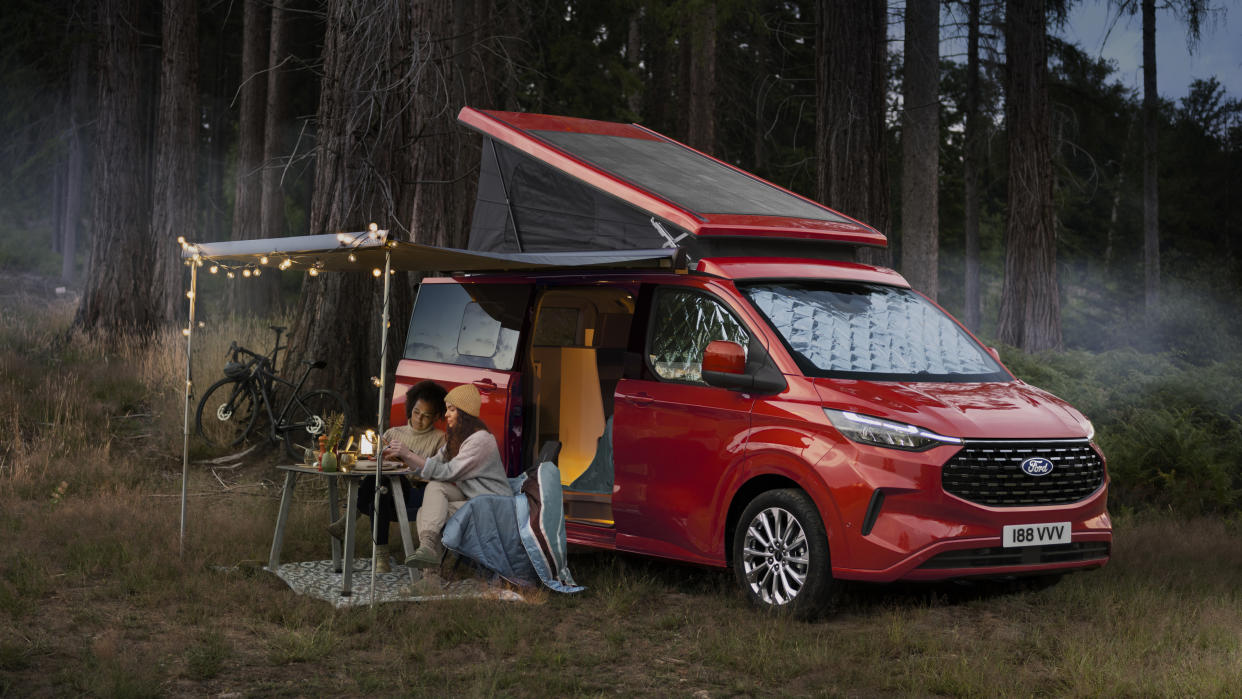  The Ford Nugget campervan in red, with people dining outside by some trees in a field 